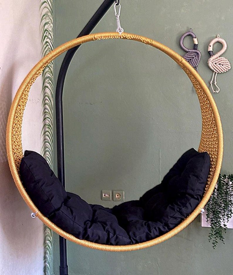 May include: A round, gold-colored hanging chair with a black cushion. The chair is made of woven rope and has a simple, modern design.