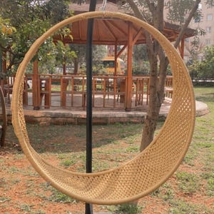 May include: A woven wicker hanging chair with a black metal stand. The chair is hanging from a chain and is suspended in the air. The chair is in a garden setting with green grass and trees in the background.
