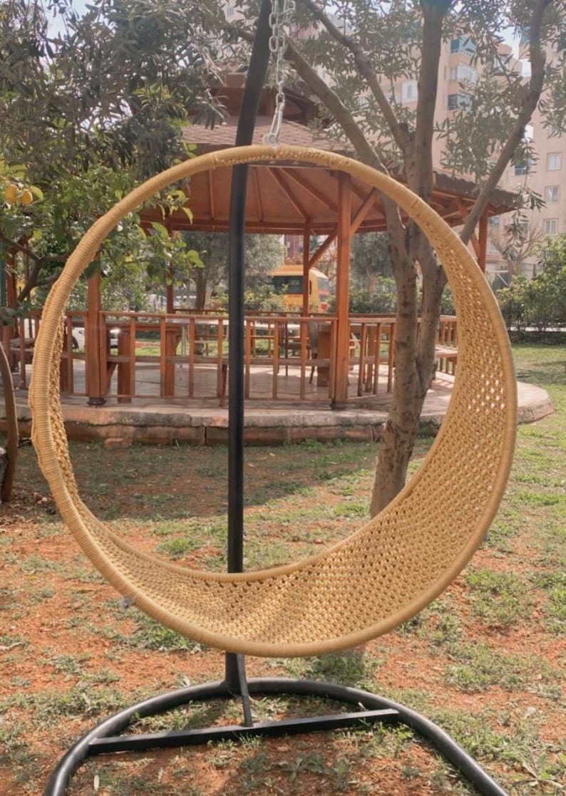 May include: A woven wicker hanging chair with a black metal stand. The chair is hanging from a chain and is suspended in the air. The chair is in a garden setting with green grass and trees in the background.
