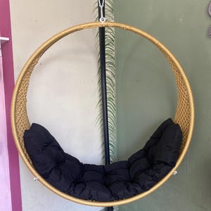 May include: A round, gold-colored hanging chair with a black cushion. The chair is made of woven rope and has a metal frame.