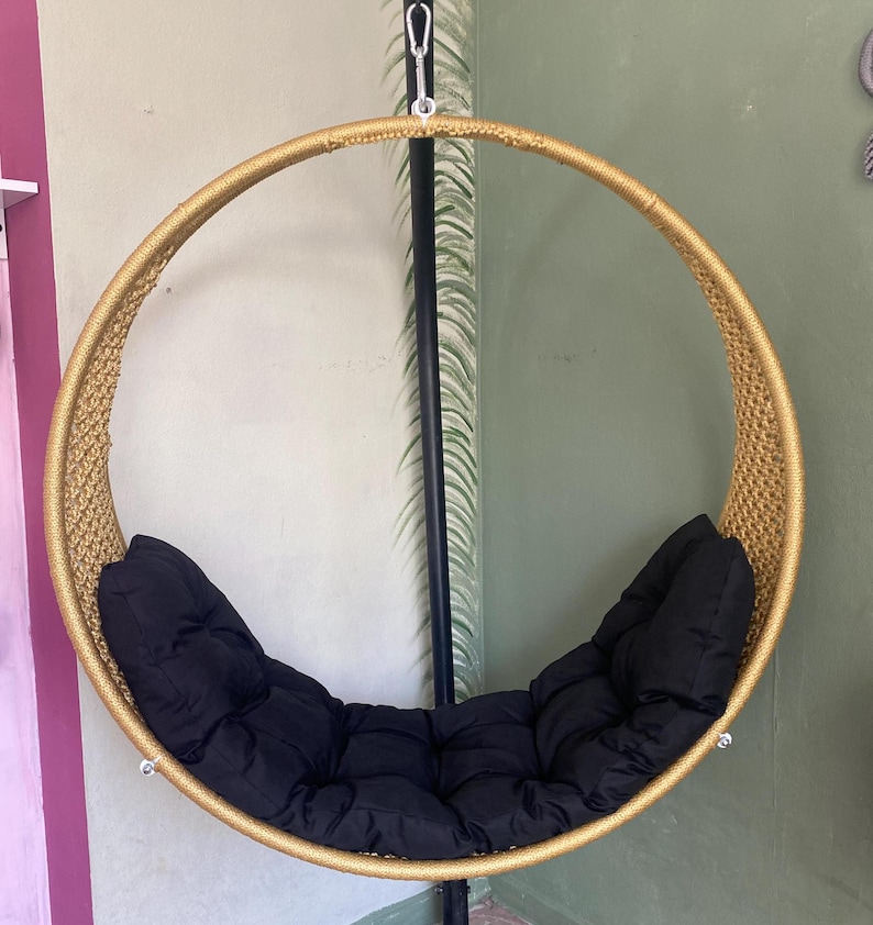 May include: A round, gold-colored hanging chair with a black cushion. The chair is made of woven rope and has a metal frame.