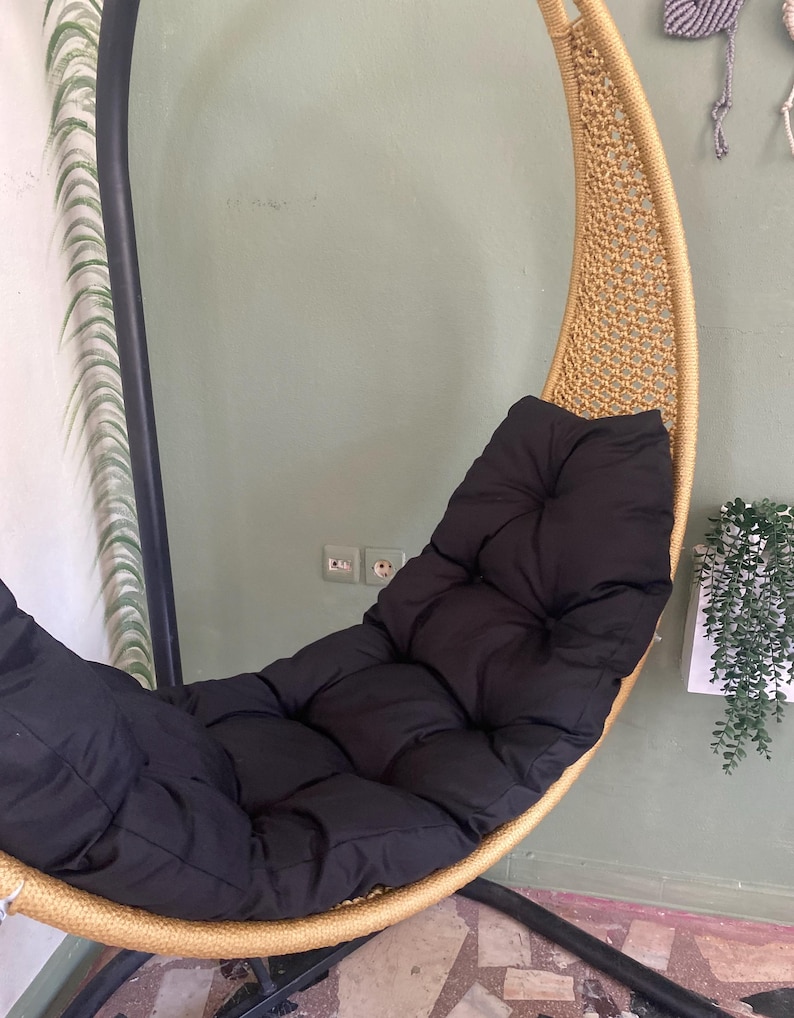 May include: A hanging chair with a black cushion. The chair is made of woven rope and has a gold finish. The chair is suspended from a black metal stand.