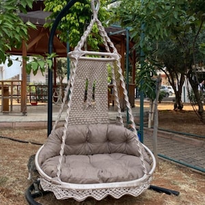 Puede incluir: Una silla hamaca colgante beige con un diseño de cuerda gruesa y un cojín marrón. La silla está colgada de un soporte de metal negro en una zona de césped.