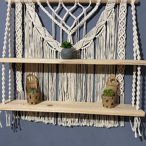 Puede incluir: Un tapiz de macramé de pared con dos estantes de madera. El macramé está hecho de cordón de algodón blanco y tiene un patrón geométrico. Los estantes están hechos de madera natural y son perfectos para exhibir plantas pequeñas u otros artículos decorativos.