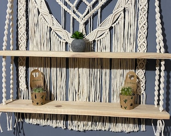 May include: A macrame wall hanging with two wooden shelves. The macrame is made of white cotton cord and has a geometric pattern. The shelves are made of natural wood and are ideal for displaying small plants or other decorative items.
