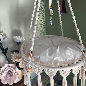 Puede incluir: Una hamaca para gatos de macramé blanco con un cojín blanco y cuentas de colores que cuelgan de la parte superior. La hamaca está suspendida del techo y tiene borlas que cuelgan de la parte inferior.