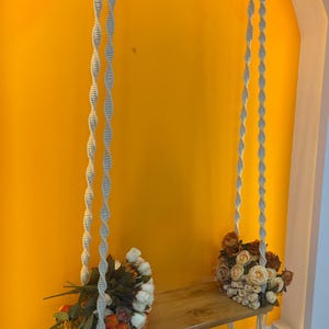 May include: A wooden swing seat suspended by two long, off-white macrame ropes featuring a spiral knot pattern. The seat is decorated on both ends with clusters of artificial flowers in shades of white, cream, orange, and deep brown.