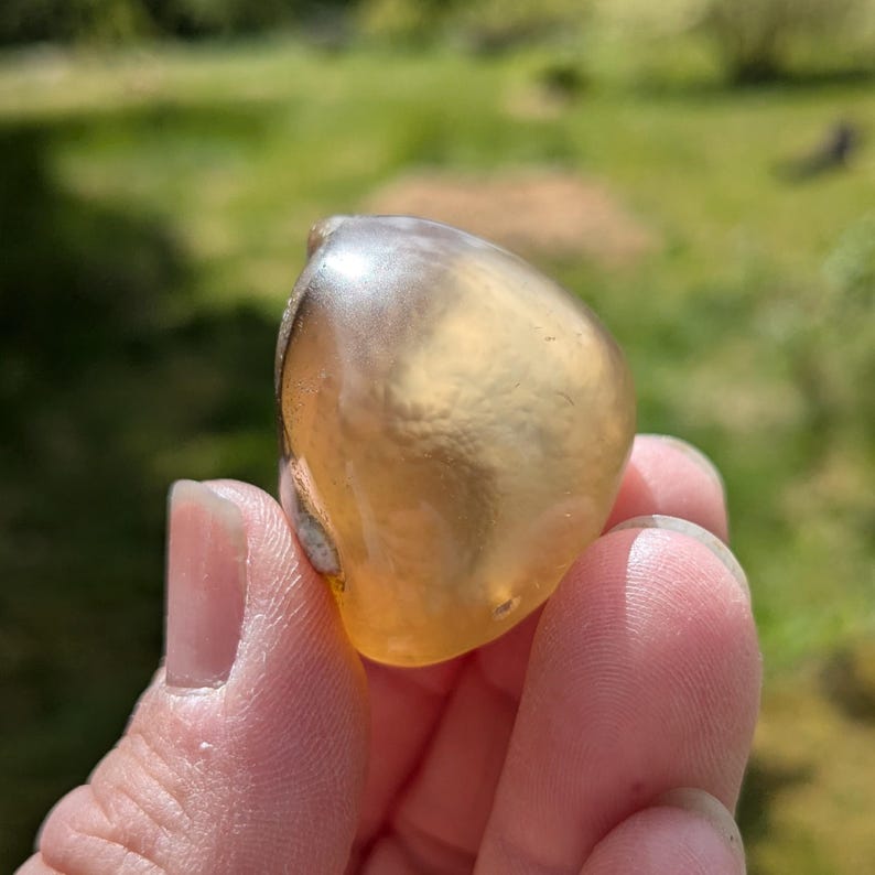 May include: A translucent, amber-colored stone with a smooth, rounded shape. The stone has a glossy surface and appears to be held in a hand, with a blurred green background.