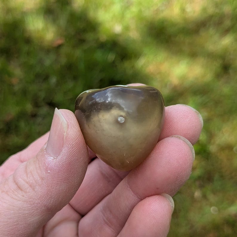 May include: A heart-shaped, polished stone with a smooth, glossy surface. The stone is a mix of brown and tan colors, with a small, light-colored inclusion near the center. The stone is held in a hand against a blurred green background.