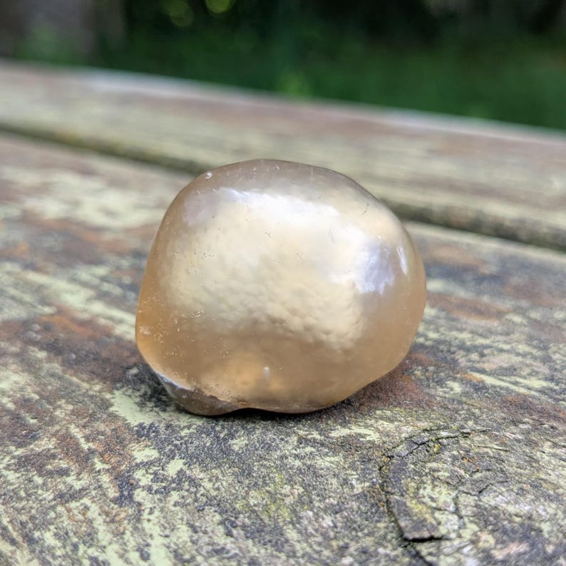 May include: A smooth, rounded, amber-colored stone with a glossy surface. The stone is resting on a weathered, wooden surface, with a blurred green background. The stone's shape is irregular, and it appears to be naturally formed.
