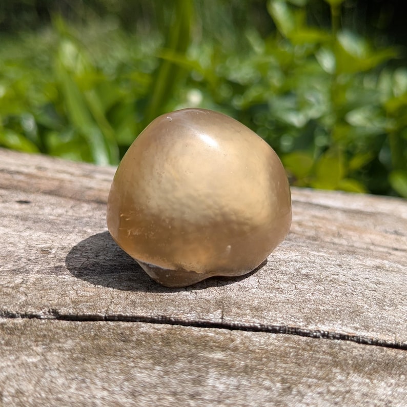 May include: A smooth, golden-colored, rounded stone with a polished surface. The stone is resting on a weathered, gray wooden surface, with a blurred green background of foliage.