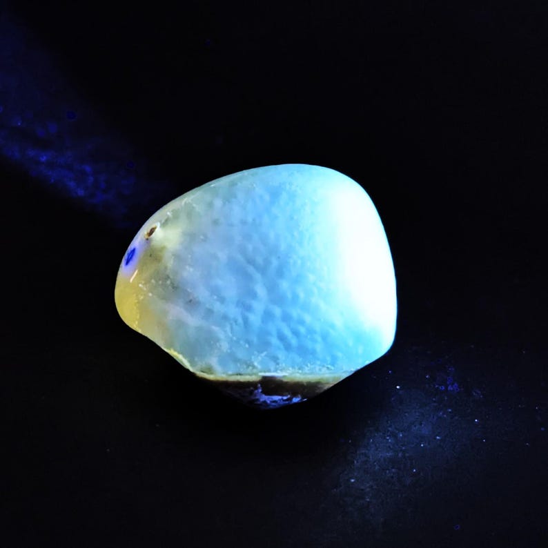 May include: A glowing, light blue gemstone with a smooth, rounded shape. The stone has a textured surface and is set against a black background. The gem appears to be illuminated from within, creating a soft, ethereal glow.