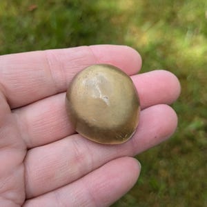 May include: A smooth, oval-shaped, golden-colored stone. The stone has a polished surface with subtle variations in color and texture. The object is held in a hand against a blurred green background.
