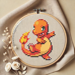 Könnte beinhalten: Kreuzstich-Stickerei eines Charmander-Charakters in einem Holzreifen. Charmander ist in Orange-, Rot- und Gelbtönen mit grünen Augen dargestellt. Der Hintergrund ist ein neutraler beigefarbener Stoff. Der Text "@dewdropdaggerco" ist sichtbar.