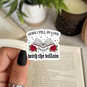 May include: A white die-cut sticker featuring an open book illustration with red roses and the black text "OOPS I FELL IN LOVE with the villain" surrounded by small black stars. The sticker is held against an open book with visible text.