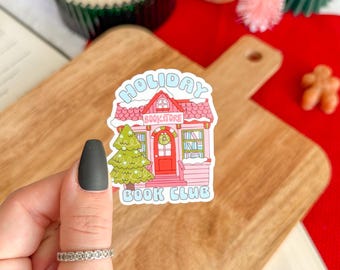 May include: A colorful sticker with the text "HOLIDAY BOOKSTORE BOOK CLUB" is held in front of a wooden cutting board. The sticker features a pink building with a green Christmas tree and a red door. Other holiday decorations are in the background.