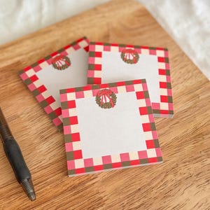May include: Three square notepads with a red and pink checkered border and a small wreath design. A black pen rests on a wooden cutting board. A small potted plant and a red candle are in the background.