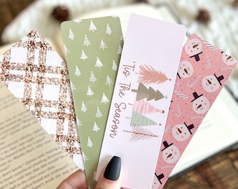 May include: Four Christmas-themed bookmarks with various designs. One has a plaid pattern, another features small Christmas trees, a third says "'Tis The Season" with trees, and the last has snowmen on a pink background. The bookmarks are held over an open book.