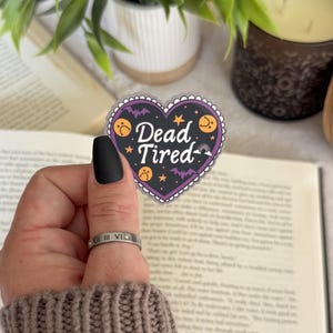 May include: A heart-shaped sticker with the words "Dead Tired" in a black font, surrounded by bats, moons, and pumpkins. The sticker is purple, black, and orange. The sticker is held in front of an open book, a candle, and a potted plant.
