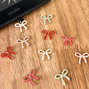 May include: A collection of small metal bow charms in various colors including red, pink, white, and light green, arranged near a black e-reader displaying the text ONE MORE CHAPTER with star graphics on its screen.