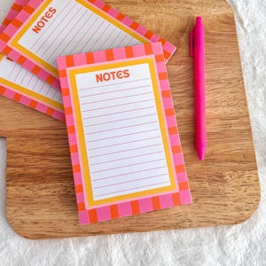 May include: A stack of rectangular note pads with a pink and orange checkered border and the word NOTES printed in orange at the top. Each pad features horizontal lines for writing. A bright pink retractable pen is placed next to the note pads.