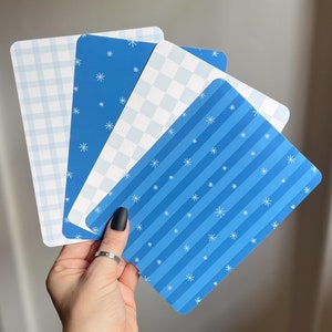 May include: Four blue and white patterned paper cards with snowflake designs. The cards are held in a hand.