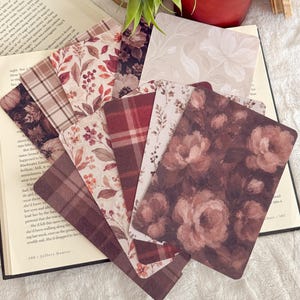 May include: A collection of rectangular bookmarks featuring various patterns including dark floral prints, red and brown plaid, and botanical illustrations. The bookmarks are fanned out over an open book, with a red candle and a green plant visible nearby.