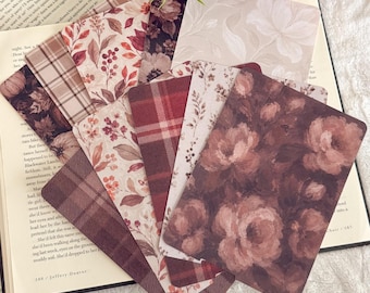 May include: A collection of rectangular bookmarks featuring various patterns including dark floral prints, red and brown plaid, and botanical illustrations with leaves and berries, fanned out over an open book with visible text.