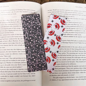 May include: Two bookmarks with different designs. One bookmark features a black and white pattern of skeletons with pink accents. The other bookmark features a pink and white pattern of fangs and spiders. Both bookmarks have a black background.