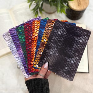 May include: A set of rectangular cards featuring a repeating dragon scale pattern in various colors including black, orange, blue, red, green, purple, and light gray. The cards are arranged in a fan shape, displaying the textured, overlapping scale design.