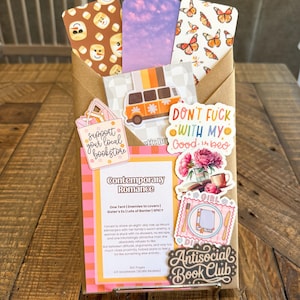 May include: A brown paper envelope containing three bookmarks with snowman, purple sky, and butterfly patterns, decorated with stickers including a van, a bookstore sign, a romance book summary card, floral art, and an Antisocial Book Club label.