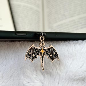 May include: A gold-colored dragon charm with black detailing and a yellow starburst center. The dragon has outstretched wings with intricate patterns and is attached to a black phone. The charm is set against a soft, white background.