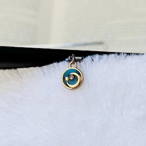 May include: A small, round pendant with a gold-colored frame and a teal enamel center. The pendant features a swirling design and a small, clear crystal. The pendant is suspended from a silver-colored bail.