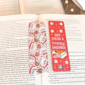 May include: Two red and white bookmark designs with a plaid background. The bookmark on the left has a white background with a pattern of Santa Claus and gingerbread cookies. The bookmark on the right has a red background with white snowflakes and the text "Hot Cocoa & Happy Endings". Both bookmarks have the text "Little Turkey Shoppe" in the bottom right corner.