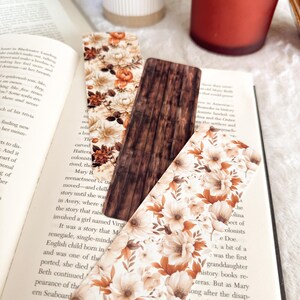 Op de afbeelding: Drie decoratieve bladwijzers liggen op een open boek. Twee hebben een bloemenpatroon in cr&egrave;me, bruin en oranje. De middelste bladwijzer heeft een houtnerf ontwerp. De pagina's van het boek zijn zichtbaar.