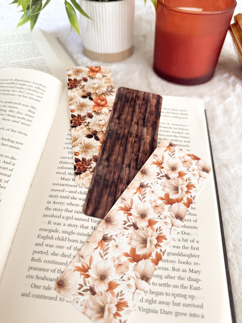 Op de afbeelding: Drie decoratieve bladwijzers liggen op een open boek. Twee hebben een bloemenpatroon in cr&egrave;me, bruin en oranje. De middelste bladwijzer heeft een houtnerf ontwerp. De pagina's van het boek zijn zichtbaar.