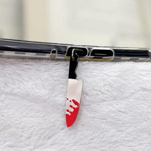 May include: A black and white phone case with a small, dangling knife charm. The knife charm has a white blade with red splatters, and a black handle. The phone case is resting on a white, fluffy surface.