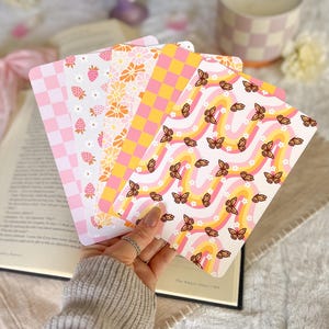 May include: A set of five rectangular bookmarks featuring various patterns including a pink and white checkerboard, pink strawberries with white flowers, orange and white daisies, a yellow and pink checkerboard, and brown butterflies on wavy lines.
