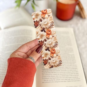 Op de afbeelding: Een rechthoekige bloemenbladwijzer met een patroon van cr&egrave;me, oranje en bruine bloemen. De bladwijzer wordt boven een open boek gehouden. De achtergrond is wazig en bevat een kaars en groen.