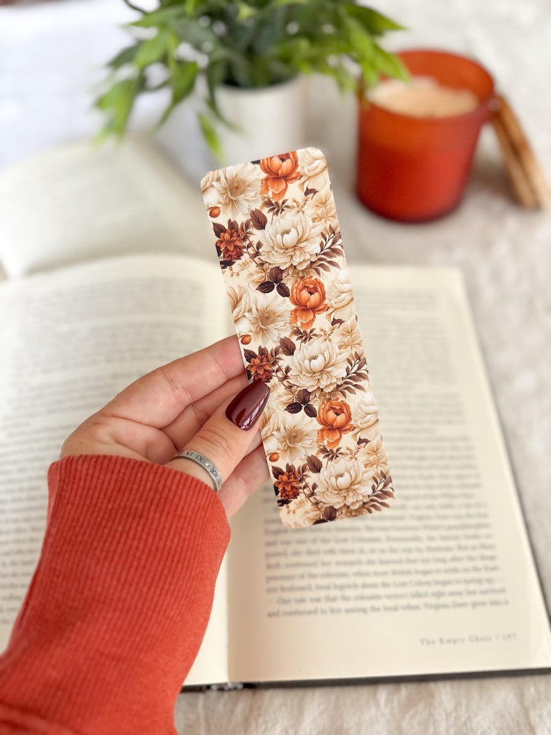 Op de afbeelding: Een rechthoekige bloemenbladwijzer met een patroon van cr&egrave;me, oranje en bruine bloemen. De bladwijzer wordt boven een open boek gehouden. De achtergrond is wazig en bevat een kaars en groen.