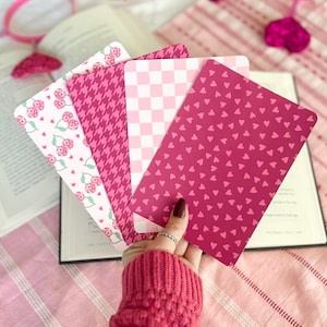 May include: Four pink patterned paper cards with different designs. The cards are stacked on top of each other and are being held by a hand wearing a pink sweater.
