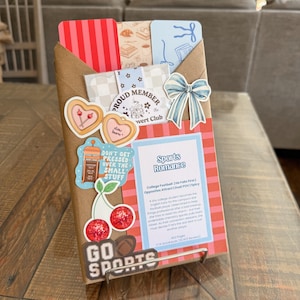 May include: A wrapped book with decorative stickers and a bow. The book is titled "Sports Romance" and has a pink and white striped cover. Stickers include cherries, hearts, and the words "Go Sports".