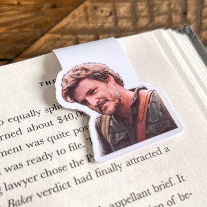 May include: A white bookmark with a photo of a man with brown hair and a beard. The man is wearing a brown leather jacket and a white shirt. The bookmark is on a book with the text "THF greed to equally split and earned about $40,00 settlement was quite pos ur and was ready to file. 7 young lawyer chose the Pr Baker verdict had finally attracted a appellant brief. It of be-" visible.