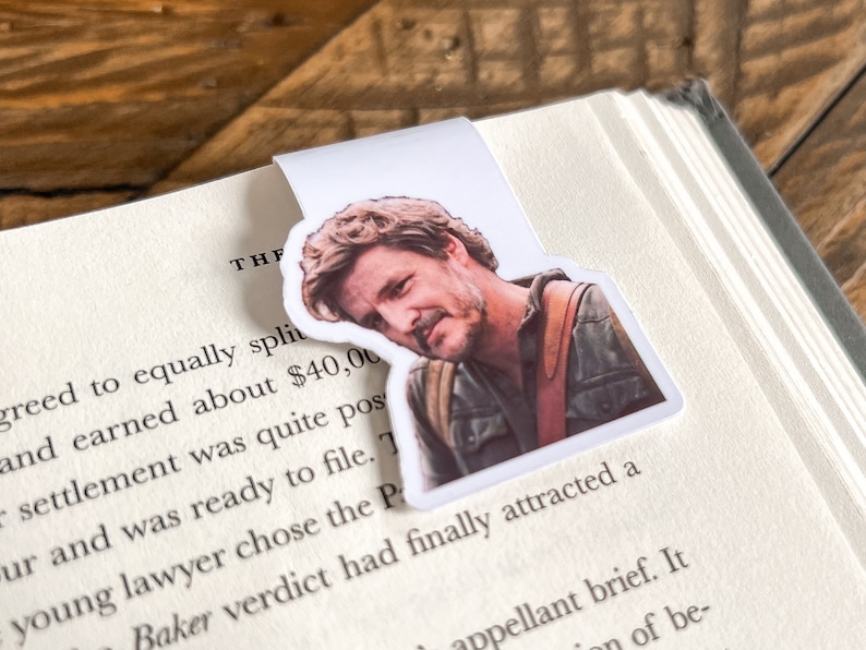 May include: A white bookmark with a photo of a man with brown hair and a beard. The man is wearing a brown leather jacket and a white shirt. The bookmark is on a book with the text "THF greed to equally split and earned about $40,00 settlement was quite pos ur and was ready to file. 7 young lawyer chose the Pr Baker verdict had finally attracted a appellant brief. It of be-" visible.