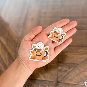 May include: Two Halloween-themed stickers featuring a ghost reading inside a pumpkin mug. The mug is orange with a black handle and a carved jack-o'-lantern face. The stickers also include fall leaves and small pumpkins.