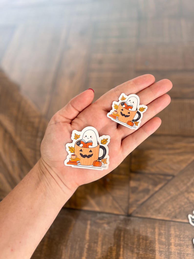 May include: Two Halloween-themed stickers featuring a ghost reading inside a pumpkin mug. The mug is orange with a black handle and a carved jack-o'-lantern face. The stickers also include fall leaves and small pumpkins.