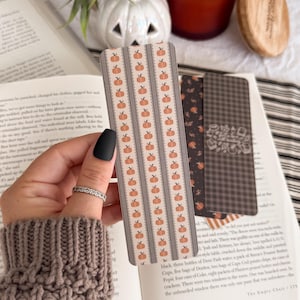 May include: A rectangular bookmark featuring a repeating pattern of small orange pumpkins arranged in vertical columns separated by thin, dark gray lace-like borders on a light beige background, with rounded corners and a smooth finish.