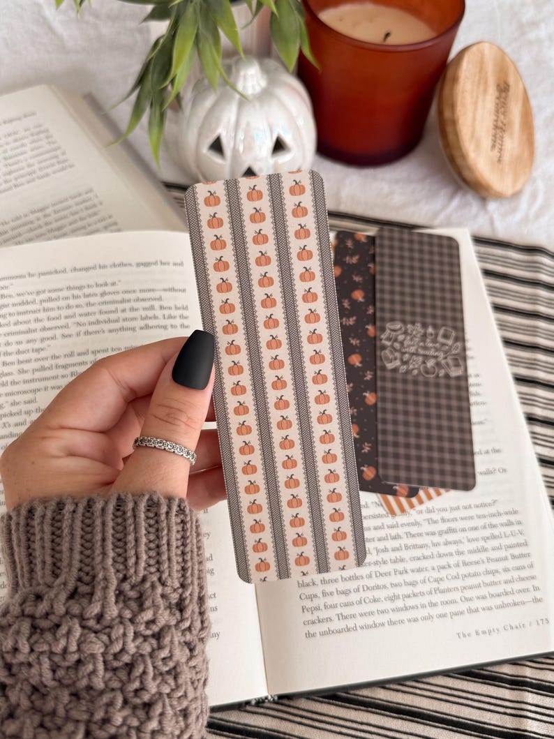 May include: A rectangular bookmark featuring a repeating pattern of small orange pumpkins arranged in vertical columns separated by thin, dark gray lace-like borders on a light beige background, with rounded corners and a smooth finish.