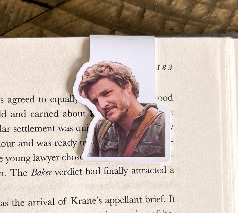 May include: A close-up of a bookmark featuring a man with brown hair and a beard. He is wearing a brown jacket and a white shirt. The bookmark is placed on a page of a book with the number 183 printed on it.