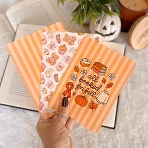 May include: Three rectangular cards with rounded corners featuring autumn-themed designs. One card displays orange stripes, a pumpkin, a sweater, books, and the text "all booked for fall." Another card features a pattern of teddy bears and bows.